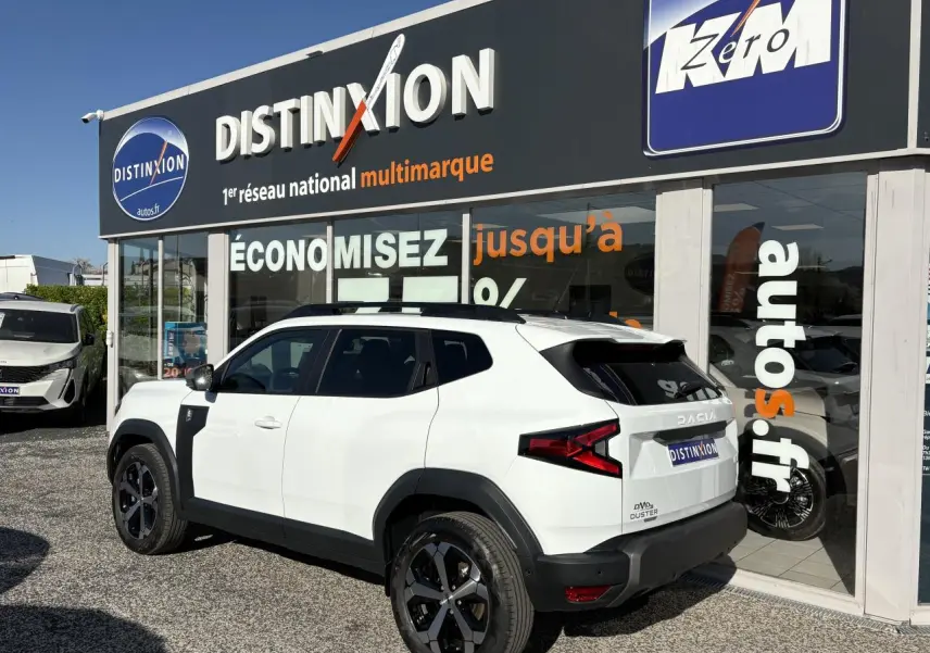 Vue 3/4 arrière droite d'un Dacia Duster blanc métallisé avec barres de toit noires devant une concession Distinxion.