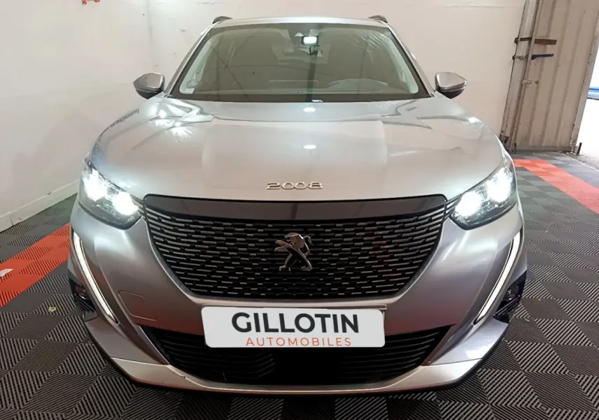 Vue frontale d'un Peugeot 2008 gris clair avec phares allumés et calandre noire distinctive dans un garage.