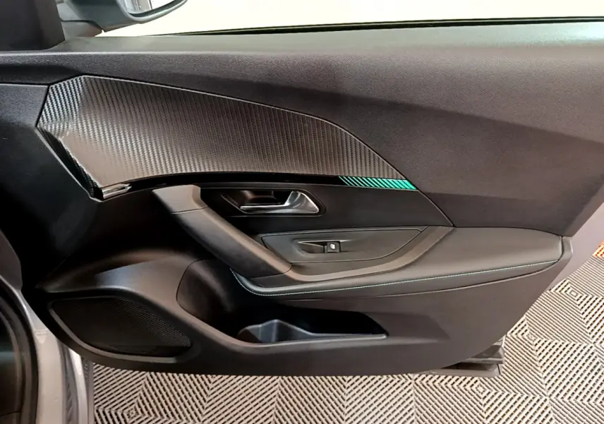 Vue intérieure côté gauche de la porte d'une Peugeot 2008 gris clair avec insert imitation carbone et surpiqûres bleues.