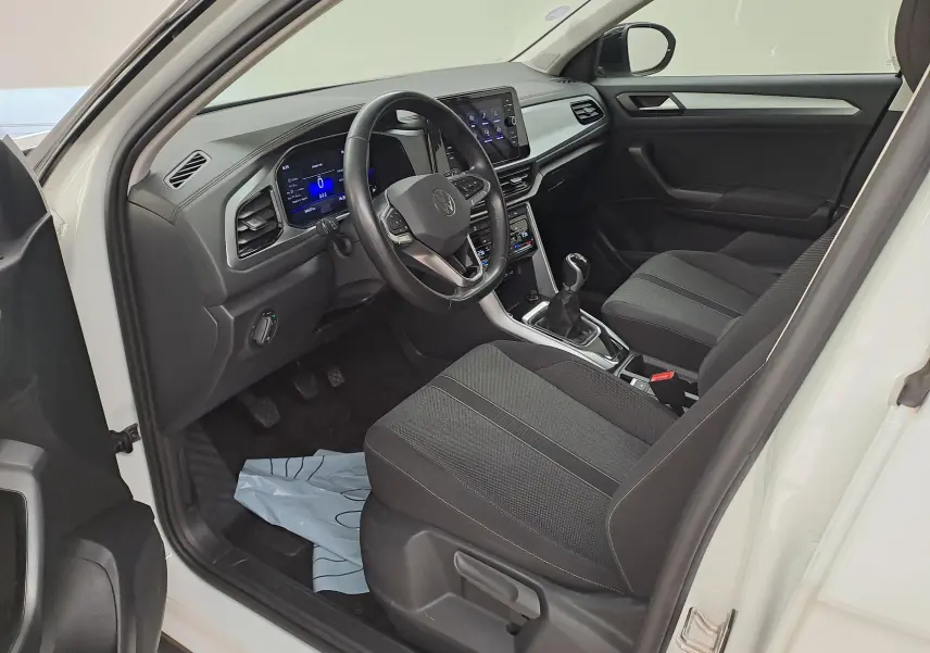 Vue intérieure côté conducteur du Volkswagen T-Roc blanc toit noir, montrant le tableau de bord numérique et la boîte manuelle.