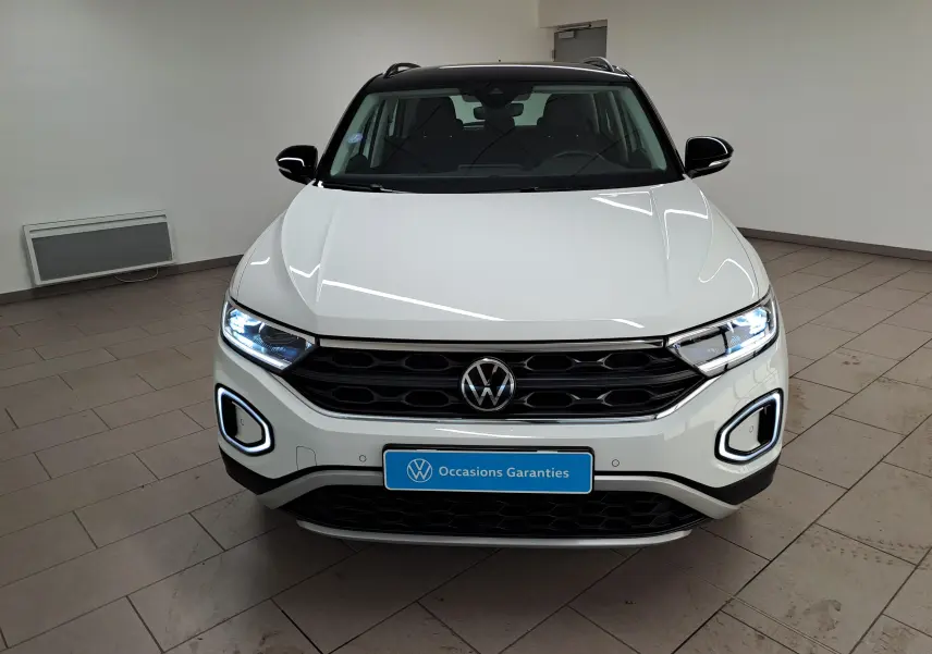 Vue avant d’un Volkswagen T-Roc blanc pur avec toit noir, phares allumés et calandre noire dans un showroom.