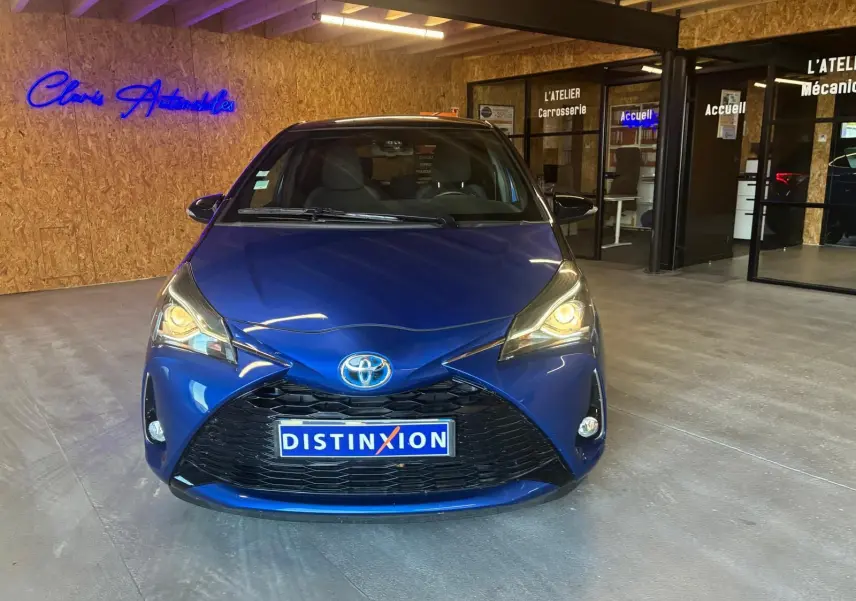 Toyota Yaris hybride bleu vue de face en intérieur, phares allumés et plaque Distinxion visible.