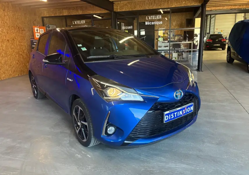 Toyota Yaris hybride bleu vue 3/4 avant droit dans un garage avec phares allumés et jantes noires.