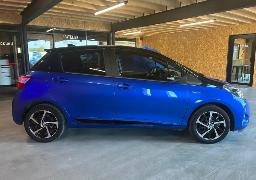 Profil côté gauche d'une Toyota Yaris hybride bleue 5 portes, avec jantes noires et toit noir en intérieur d'atelier.