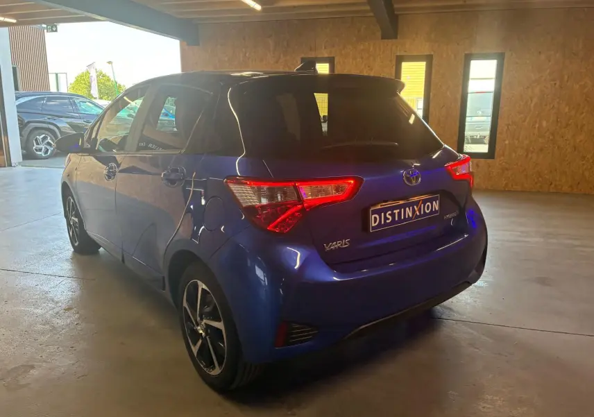 Toyota Yaris hybride bleu vue 3/4 arrière droit dans un garage, avec feux arrière allumés et jantes noires.