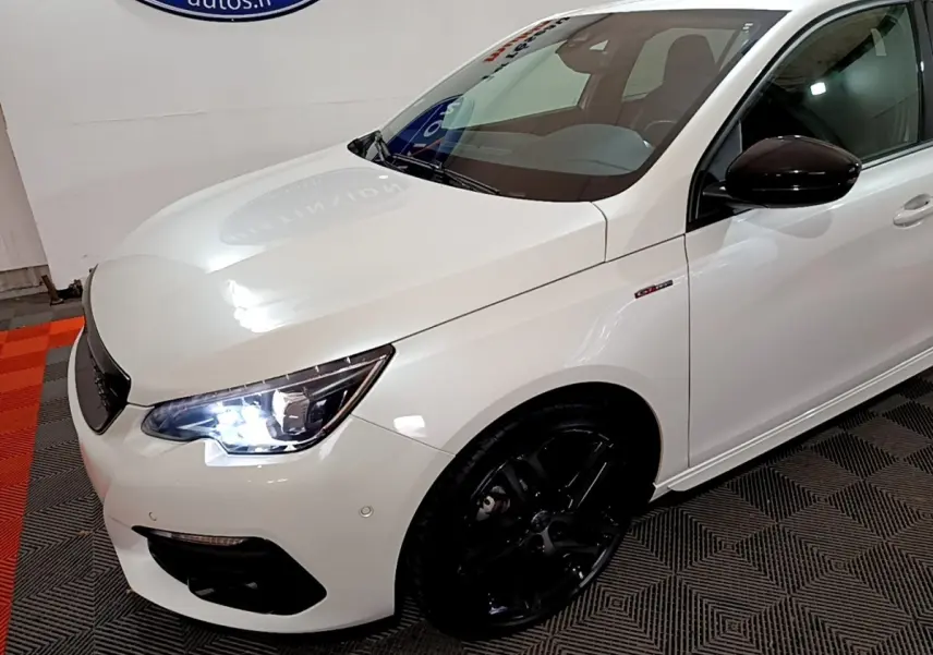 Vue 3/4 avant droit d'une Peugeot 308 blanche avec phares allumés et jantes noires brillantes.