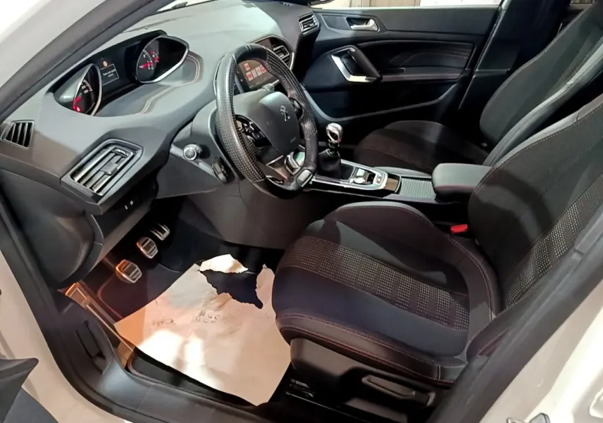 Vue intérieure côté conducteur de la Peugeot 308 GT Line blanche, montrant le volant cuir, la boîte manuelle et les sièges noirs.