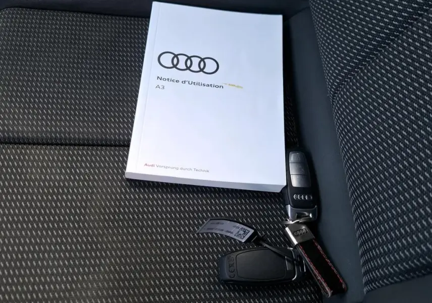 Notice d'utilisation Audi A3 posée sur un siège tissu noir à motifs avec une clé de voiture Audi à côté.