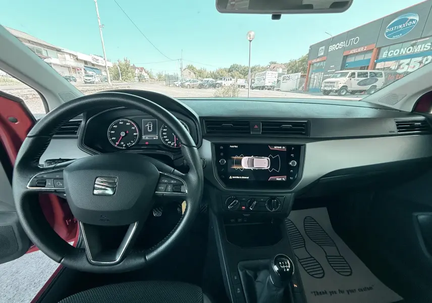 Vue intérieure centrée sur le volant cuir et tableau de bord d’une SEAT Ibiza rouge passion, avec écran tactile et boîte manuelle.