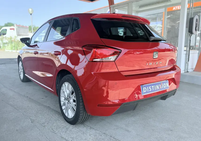 Vue 3/4 arrière droite d'une SEAT Ibiza rouge passion métal avec hayon et jantes argentées, stationnée devant un bâtiment.