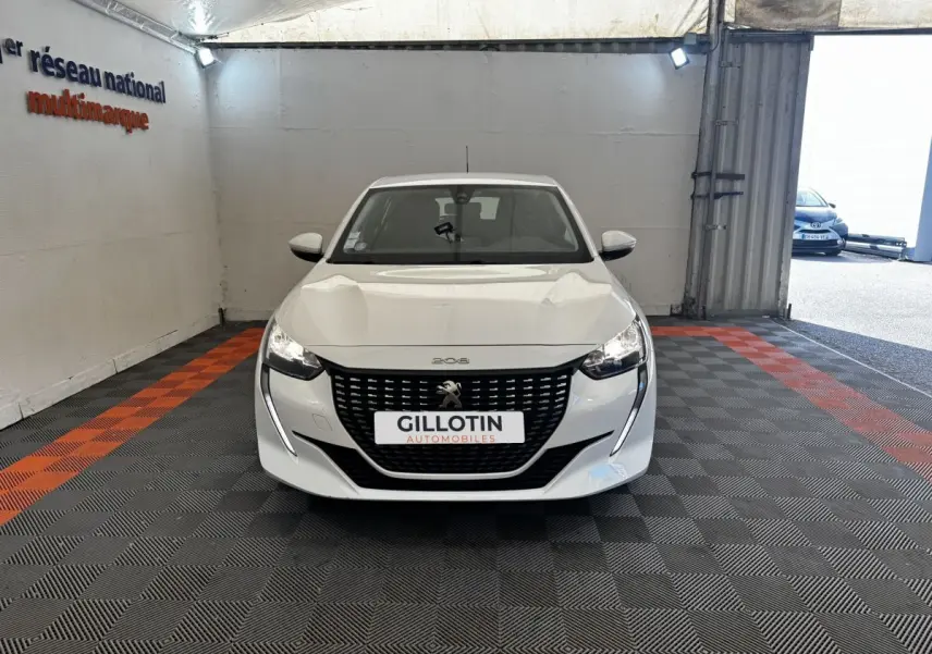 Vue avant d'une Peugeot 208 blanche stationnée en intérieur, avec calandre noire et phares allumés.