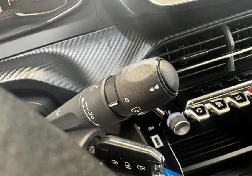 Gros plan sur la commande d’essuie-glace noire et la console centrale avec boutons et finition carbone dans l’habitacle d’une Peugeot 208 blanche.
