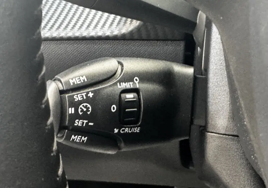 Gros plan sur la commande de régulateur de vitesse noire à gauche du volant d'une Peugeot 208 blanche.