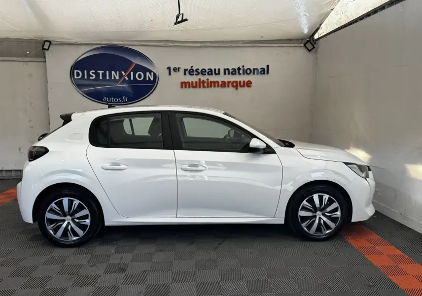 Profil latéral droit d'une Peugeot 208 blanche stationnée en intérieur, avec jantes alliage et vitres teintées.