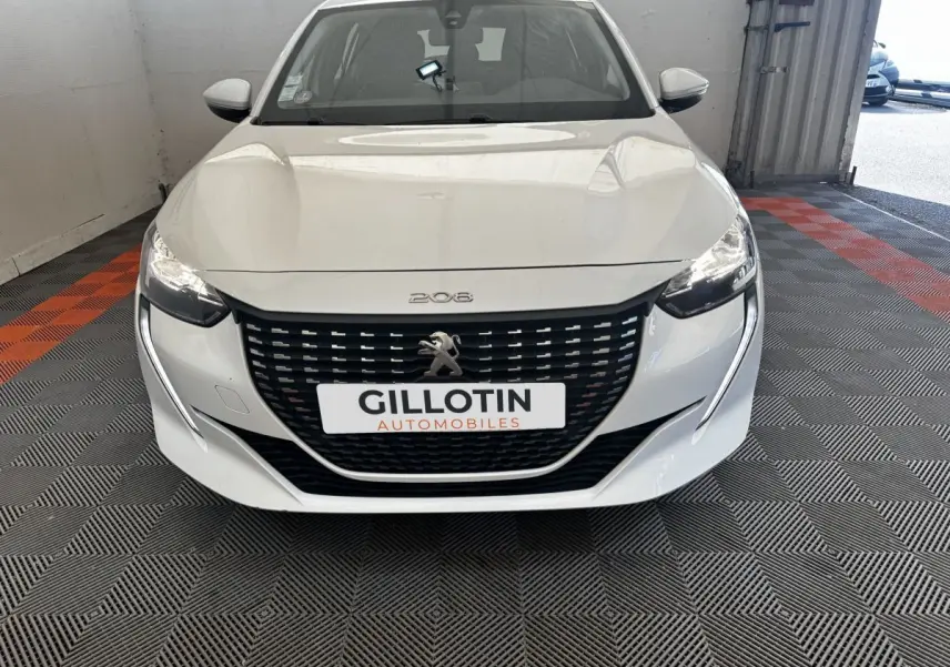 Vue avant d'une Peugeot 208 blanche avec calandre noire et phares allumés dans un garage.