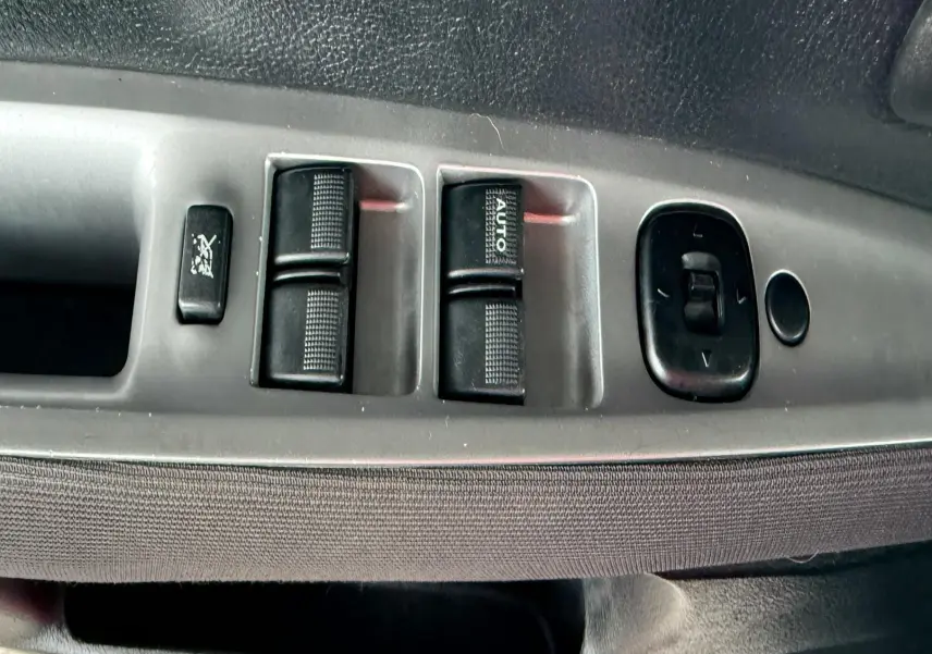 Gros plan sur les commandes des vitres électriques et rétroviseurs côté conducteur d'une Mazda2 gris 2004.