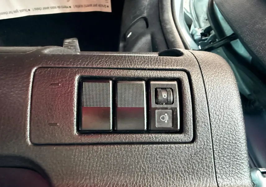 Gros plan sur les commandes de réglage des phares dans l'habitacle noir d'une Mazda2 gris 2004.