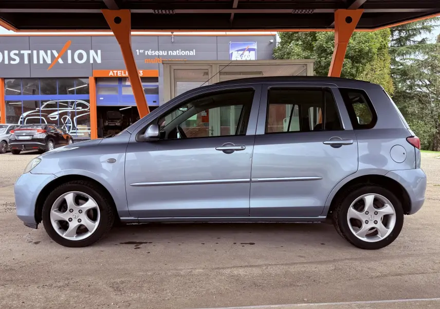 Vue latérale côté gauche d'une Mazda2 gris clair 5 portes de 2004 stationnée sous un abri orange.