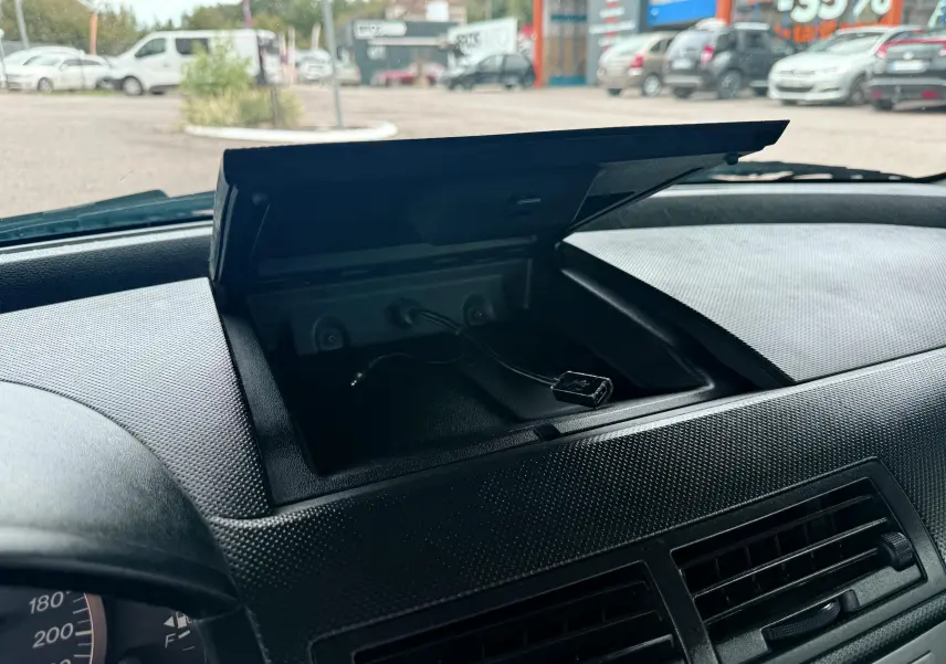 Coffre de rangement ouvert sur le tableau de bord d'une Mazda2 gris 2004, avec câble USB visible.