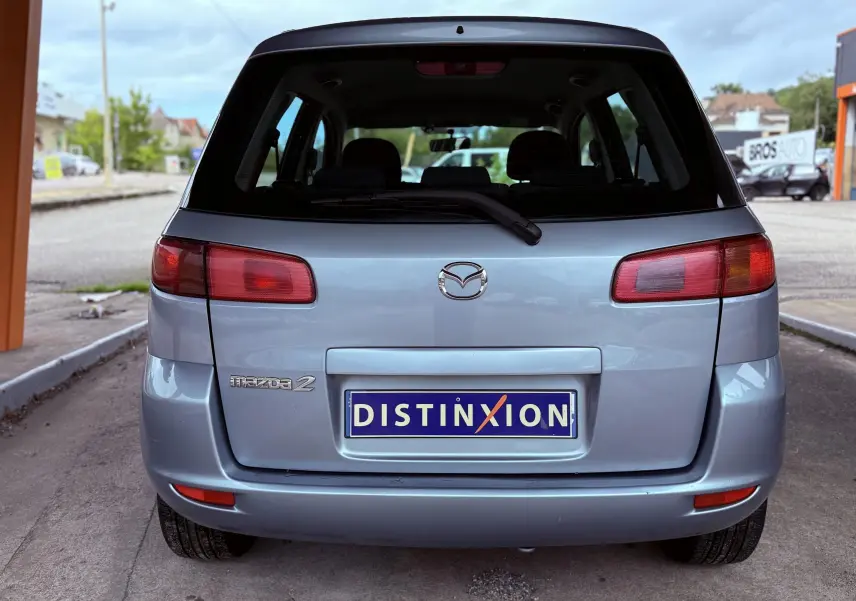 Vue arrière d'une Mazda2 gris clair de 2004 avec logo Mazda et plaque d'immatriculation Distinxion visible.
