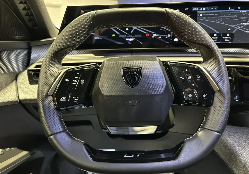 Volant cuir perforé noir avec logo Peugeot et inscription GT, vue frontale du poste de conduite du 5008 Hybrid 2025.