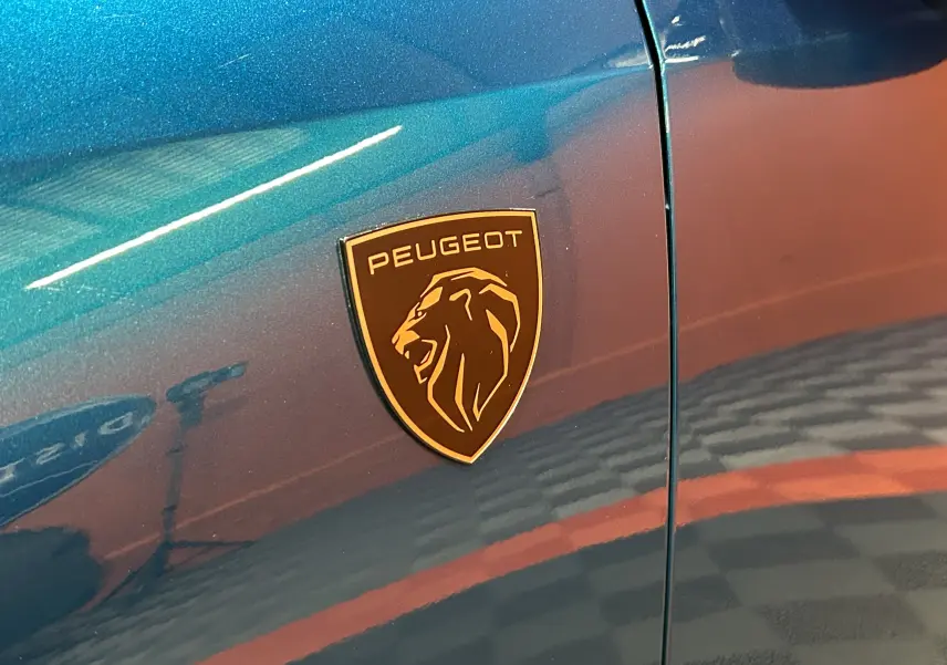 Gros plan sur l'écusson Peugeot doré sur la carrosserie bleu obsession du côté gauche du Peugeot 5008 III Hybrid 145 GT.