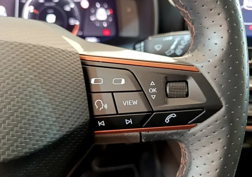 Gros plan sur les commandes noires et orange du volant en cuir perforé du CUPRA Formentor 2023, affichage digital en arrière-plan.