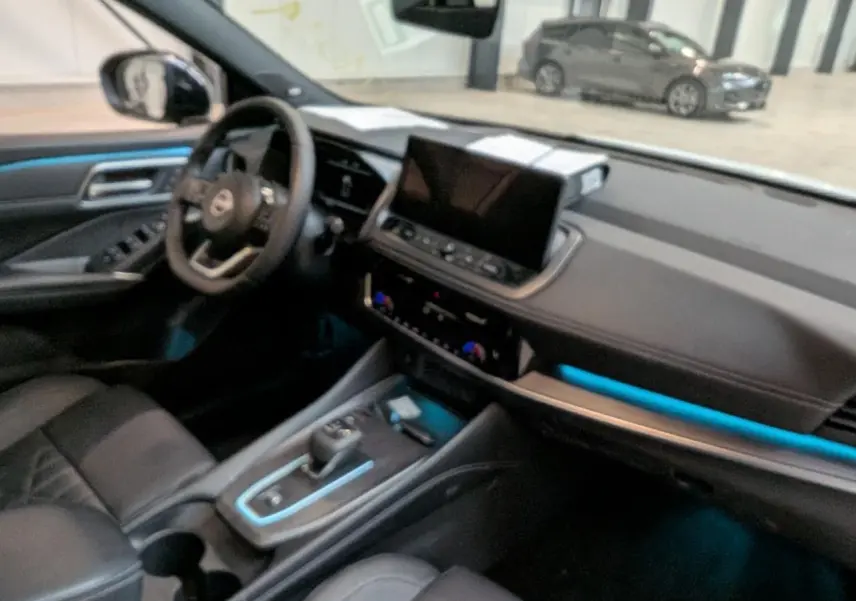 Vue intérieure côté conducteur du Nissan Qashqai 2025, sellerie noire avec éclairage d'ambiance bleu sur la console et tableau de bord.