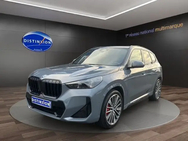 BMW X1 Storm Bay en 3/4 avant droit, jantes alliage 20'' et freins M Sport rouges visibles dans un showroom.