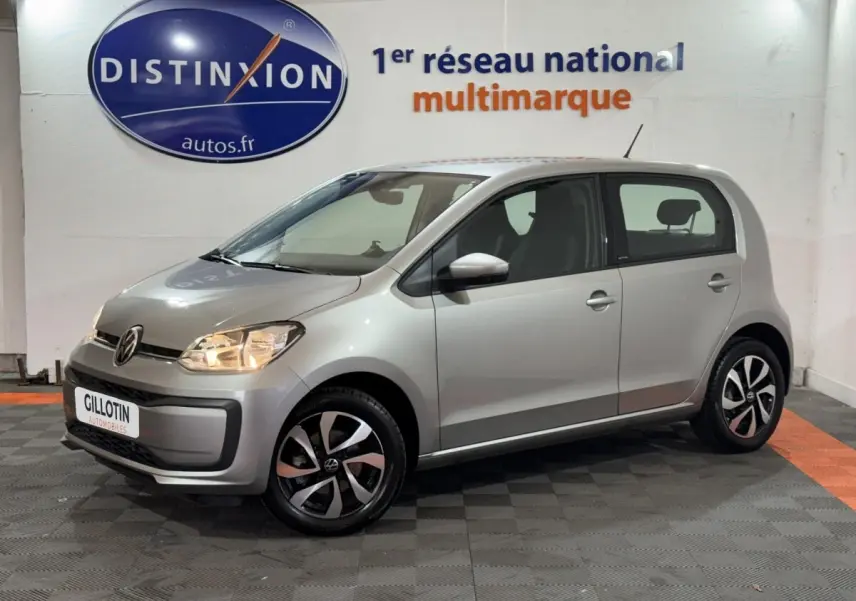 Volkswagen Up gris clair vue de profil côté droit, feux allumés, jantes noires et argentées en intérieur showroom.