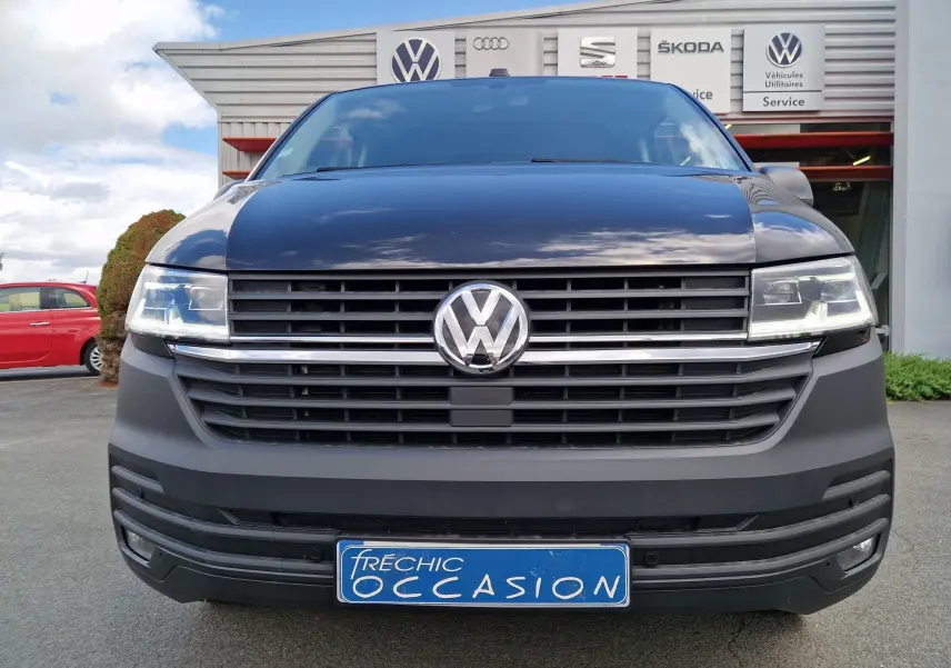 Vue frontale d'un Volkswagen Transporter noir avec projecteurs LED et calandre horizontale, devant un garage multimarque.