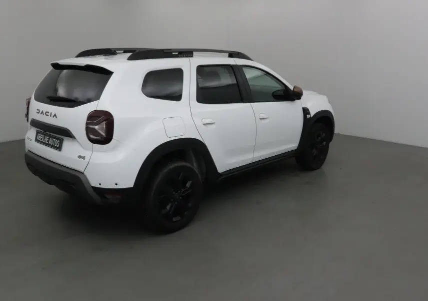 Vue 3/4 arrière droite d'un Dacia Duster blanc 4x4 avec jantes noires et barres de toit dans un studio.