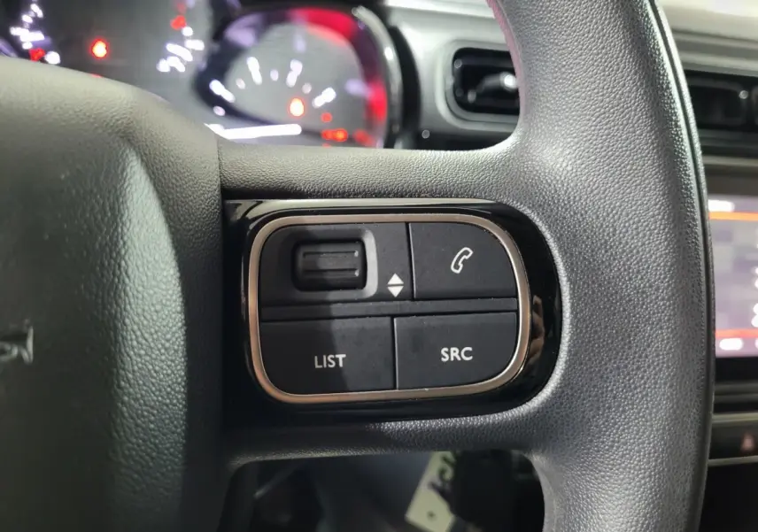 Gros plan sur les commandes gauche du volant noir de la Citroën C3 beige, avec tableau de bord et écran tactile en arrière-plan flou.