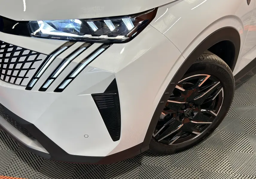Gros plan sur l’avant droit blanc Okenite du Peugeot 3008 III Hybrid 145 GT avec phares LED et jante noire brillante.