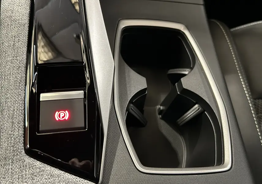 Gros plan sur la console centrale noire et grise du Peugeot 3008 Hybrid 2025 avec porte-gobelets et bouton frein à main électrique allumé.
