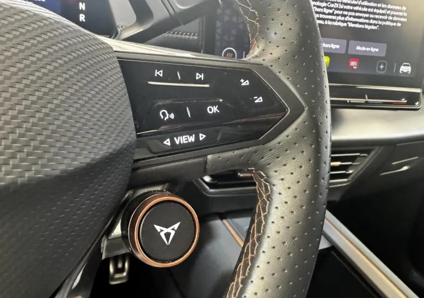 Gros plan sur le volant cuir perforé avec commandes tactiles et logo CUPRA, tableau de bord et écran tactile en arrière-plan.