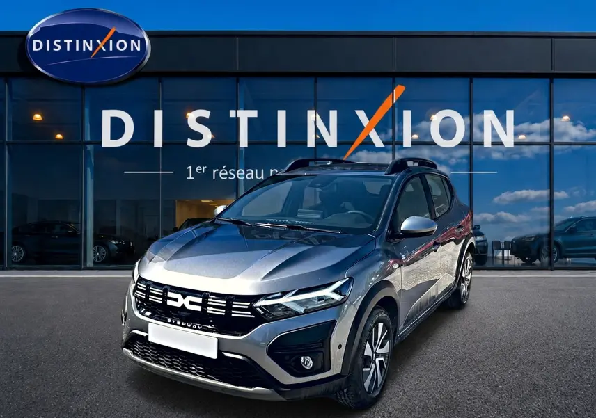 Dacia Sandero Stepway gris schiste vue 3/4 avant droit avec barres de toit devant un bâtiment Distinxion.