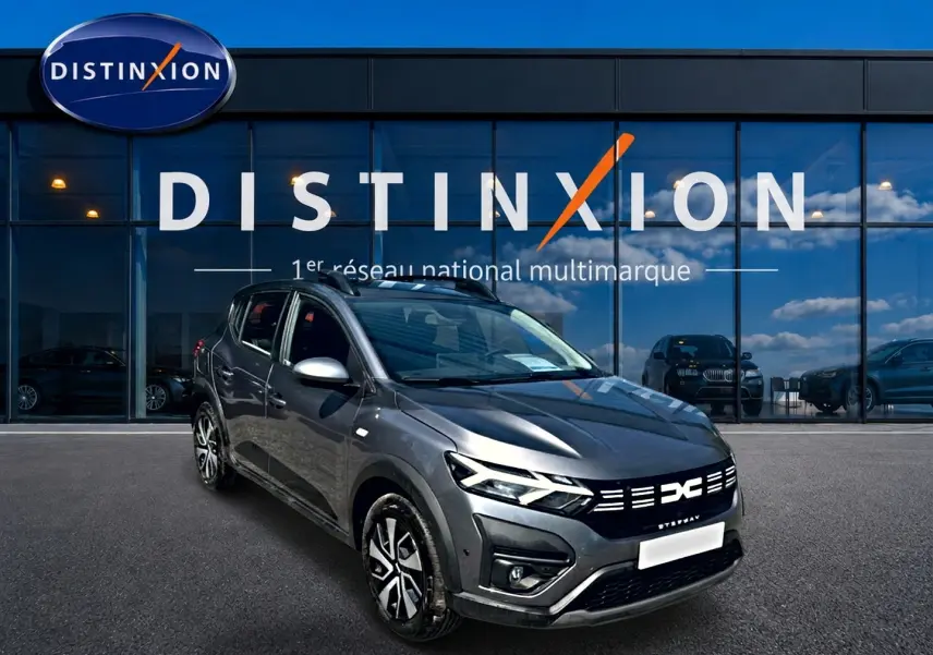 Vue 3/4 avant droit d'une Dacia Sandero Stepway gris schiste avec barres de toit et jantes distinctives devant un showroom Distinxion.