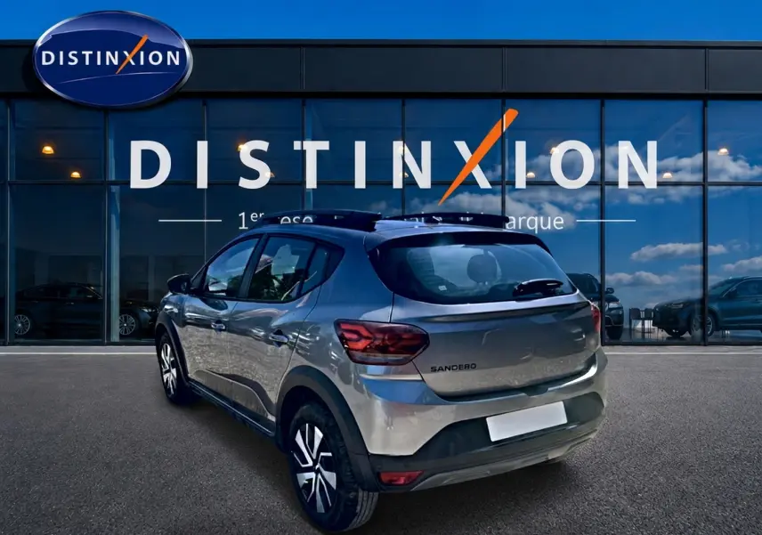 Vue 3/4 arrière droite d'une Dacia Sandero gris schiste avec barres de toit et jantes distinctives, devant un showroom Distinxion.