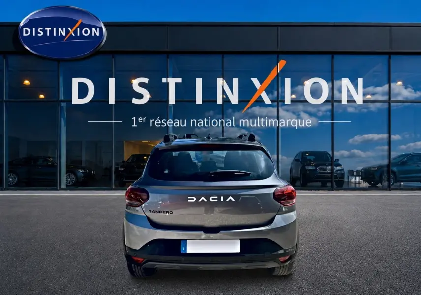 Vue arrière d'une Dacia Sandero gris schiste 2025 avec barres de toit et logo distinctif sur fond de vitrine Distinxion.