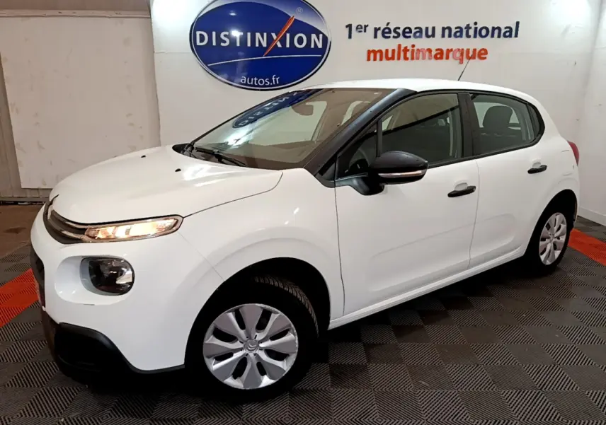 Citroën C3 blanche vue en 3/4 avant droit, avec feux allumés et rétroviseurs noirs dans un showroom.