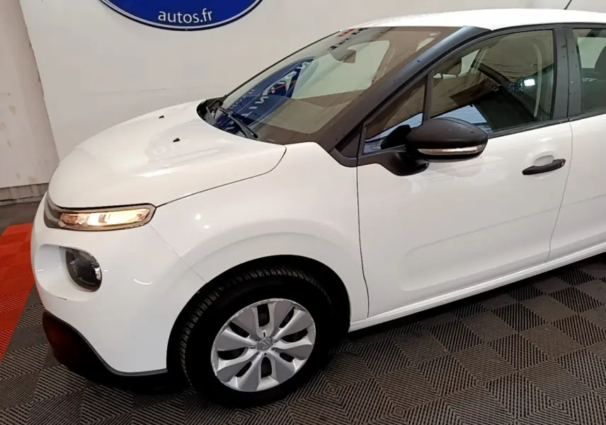 Vue 3/4 avant gauche d'une Citroën C3 blanche avec rétroviseurs noirs et jantes argentées en intérieur showroom.