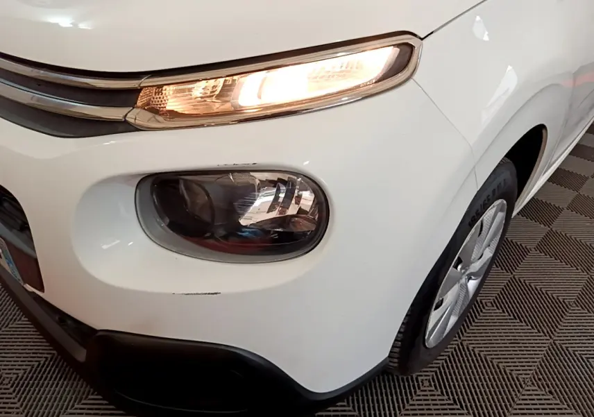 Gros plan sur l'avant droit d'une Citroën C3 blanche de 2018, mettant en valeur ses phares allumés et la roue.