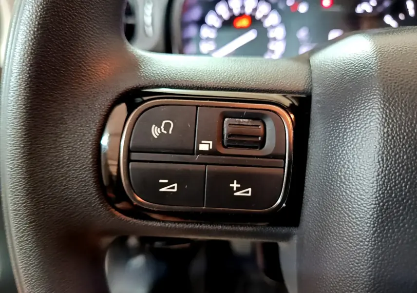 Gros plan sur les commandes multifonctions noires du volant d'une Citroën C3 beige, avec tableau de bord flou en arrière-plan.