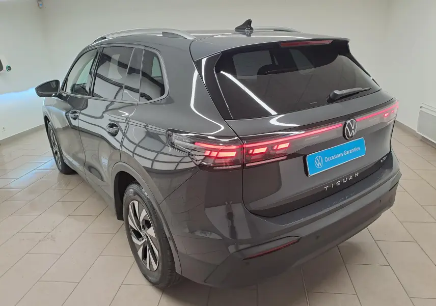 Vue 3/4 arrière droite du Volkswagen Tiguan 2024 gris Urano avec feux arrière allumés et crochet d'attelage escamotable visible.