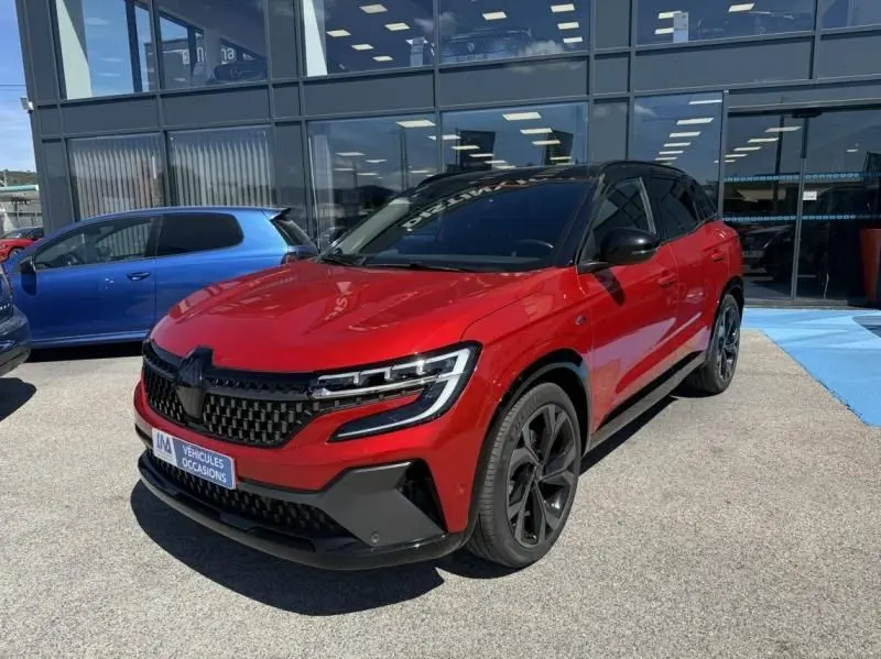 Renault Austral rouge flamme en 3/4 avant droit, avec calandre noire et jantes noires distinctives, stationné devant un bâtiment vitré.