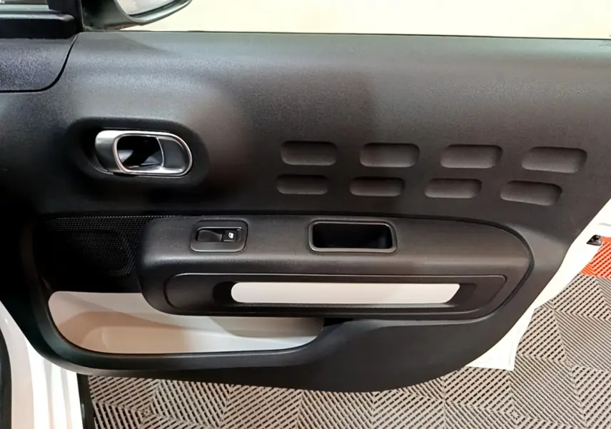 Vue rapprochée de la porte intérieure côté gauche d'une Citroën C3 blanche avec poignée argentée et panneau noir texturé.