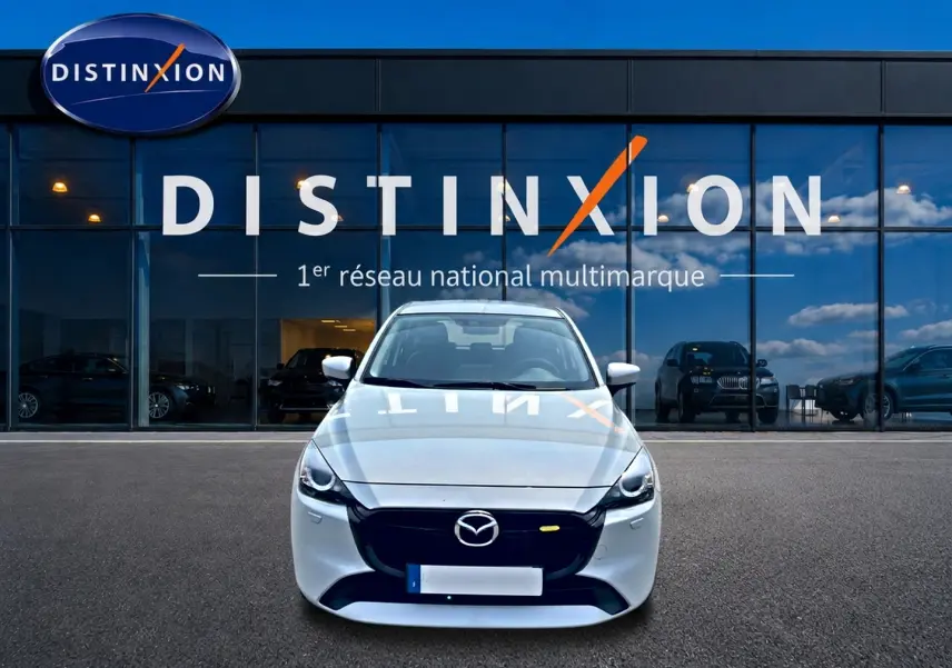 Vue frontale d'une Mazda2 blanche 2024 garée devant une vitrine avec logo Distinxion en reflet sur le capot.