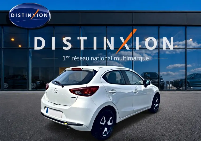 Vue 3/4 arrière droite d'une Mazda2 blanche 2024 garée devant une vitrine avec logo Distinxion.