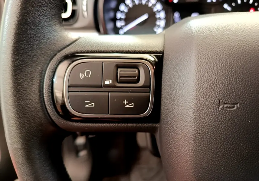 Gros plan sur les commandes gauche du volant cuir de la Citroën C3 gris foncé, avec tableau de bord flou en arrière-plan.